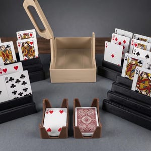 May include: A set of black tiered card holders displaying various playing cards, a beige plastic card shuffler with an open lid, and two brown plastic card trays holding a deck of cards with red patterned backs and a four of hearts.