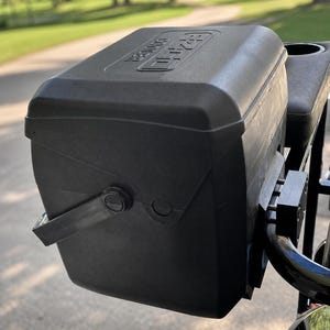 May include: A black plastic cooler mount attached to a golf cart frame. The mount features a rectangular bracket system with visible metal hardware. The text E-Z-GO COOLER MOUNT is displayed at the top, and MOUNT & HARDWARE ONLY COOLER NOT INCLUDED at the bottom.