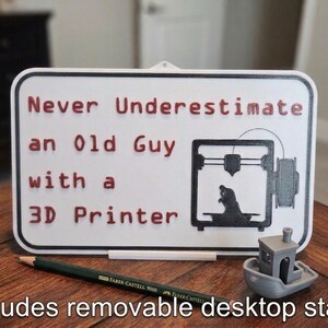 May include: A white sign with a black border and the text "Never Underestimate an Old Guy with a 3D Printer" in red. A 3D printer graphic is on the right. A small gray 3D printed boat and a pencil are on the table.