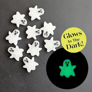 May include: White ghost-shaped decorations with black eye and mouth details are scattered on a dark surface. A circular graphic states "Glows In The Dark!" and shows a glowing green ghost. These Halloween-themed decorations are perfect for crafts.