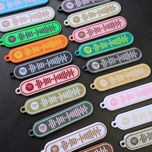 May include: A collection of colorful Spotify code keychains in various colors, including blue, green, orange, black, pink, and gold. Each keychain features a unique audio waveform design and the Spotify logo. The keychains have a small loop for attachment.
