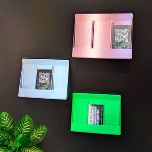 May include: Three colourful, rectangular holders in pink, light blue, and green, each with a QR code card. The pink holder has two vertical dividers. The green holder has text that reads "The Moss Forge...JUST LIKE YOU."