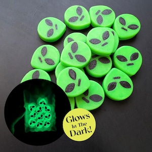 May include: A collection of green alien-shaped objects with dark gray facial features. A circular graphic shows the aliens glowing in the dark. A yellow sticker with the text "Glows In The Dark!" is also visible.