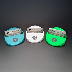 May include: Three colorful business card holders. The holders are teal, white, and green, each with a slot for cards and a wireless symbol. Each holder displays a business card with the text "The Moss Forge" and "A LITTLE wild, A LOT OF style."