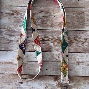 Pode incluir: Um cordão de tecido colorido com um padrão floral em vermelho, verde, amarelo e azul. O cordão tem um fecho de metal prateado na parte inferior. O fundo é uma superfície de madeira castanha clara.