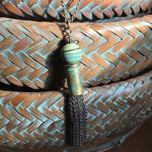 May include: A handmade necklace featuring a decorative tassel pendant. The pendant has a striped green and brown bead, a tarnished brass tube, and a chainmail fringe. The necklace chain is a bronze color.