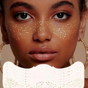 May include: A person's face with gold glitter freckles and gold hoop earrings. A white, scalloped-edge mask with gold glitter is held in front of the lower face. The skin tone is brown with full lips and dark eyes. The image is a close-up.