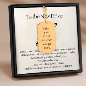 May include: A gold-colored dog tag pendant with the text "Drive safe & text me when you get there" hanging in front of a framed card. The card reads "To the New Driver" and includes a message of encouragement.