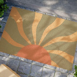 Può includere: Un tappetino per la porta con un design a raggiera in tonalità di verde, arancione e marrone. Il sole è composto da linee sottili e curve che si irradiano da un grande cerchio arancione al centro.