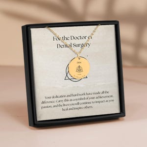 May include: A gold necklace with a round pendant that says "DDS" and has a symbol of a caduceus inside a triangle. The necklace is in a black box with a message that says "For the Doctor of Dental Surgery".