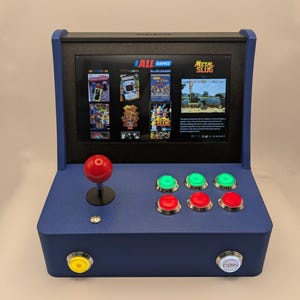 May include: A blue tabletop arcade game console with a screen displaying various game titles. It features a red joystick, green and red buttons, and a coin button. The screen displays the text "ALL GAMES" and "METAL SLUG".
