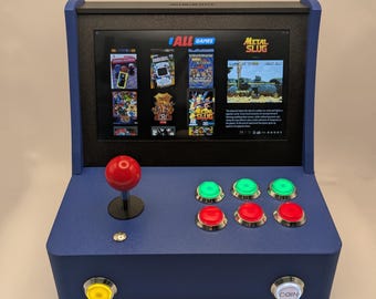 May include: A blue tabletop arcade game console with a screen displaying various game titles. It features a red joystick, green and red buttons, and a coin button. The screen displays the text "ALL GAMES" and "METAL SLUG".