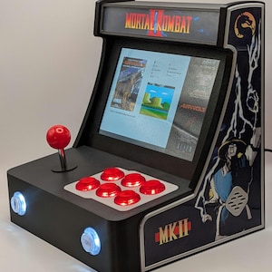 May include: A black Mortal Kombat II arcade machine with a red joystick and buttons. The screen displays game graphics. The cabinet features the game's logo and character art, with glowing coin buttons.