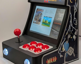 May include: A black Mortal Kombat II arcade machine with a red joystick and buttons. The cabinet features the game's logo and character art. The screen displays gameplay. The machine has two coin buttons and two lights.