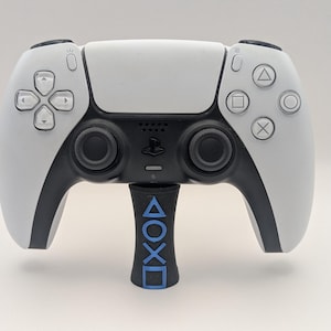 Könnte beinhalten: Weiß-schwarzer PlayStation 5-Controller mit einem schwarz-blauen Ständer. Der Ständer zeigt die PlayStation-Symbole in Blau. Der Controller hat eine weiße Frontplatte, schwarze Akzente und schwarze Analogsticks.