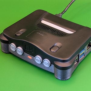 Pode incluir: Um console de jogos retrô preto com detalhes cinza, com o número "64" e vários botões redondos. O console tem um cabo de alimentação conectado e está em um fundo verde.