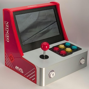 May include: A small arcade cabinet in red and silver, with a screen, joystick, and coloured buttons. The cabinet has the text "NEO-GEO SYSTEM" and "MVS" on the side, and a "COIN" button.
