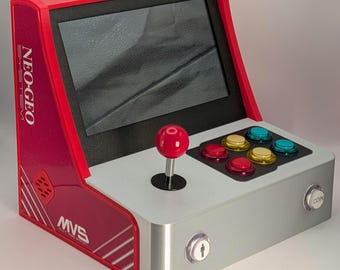 May include: A miniature arcade cabinet in red and silver, featuring a screen, joystick, and colorful buttons. The cabinet has the text "NEO-GEO SYSTEM" and "MVS" on the side, and a "COIN" button.