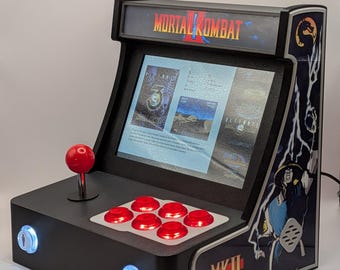 Op de afbeelding: Een tafelmodel arcadekast met Mortal Kombat II-branding, een scherm met spelmenu's, een rode joystick en zes rode actieknoppen. Het zijpaneel toont karakterillustraties en twee verlichte knoppen met het label Coin bevinden zich aan de voorzijde.
