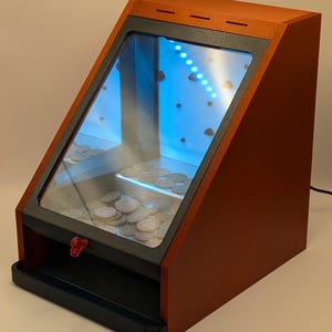 Könnte beinhalten: Ein Spielautomat aus Holz mit einer schrägen Glasfront, gefüllt mit Münzen. Das Gehäuse ist rotbraun mit schwarzem Rand und einem roten Griff. Der Innenraum verfügt über blaue LED-Leuchten.