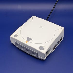Puede incluir: Una consola de videojuegos Sega Dreamcast blanca con un compartimento de disco de carga superior, un logotipo circular, un panel frontal gris con cuatro puertos para mandos y un botón de encendido con una luz indicadora naranja.
