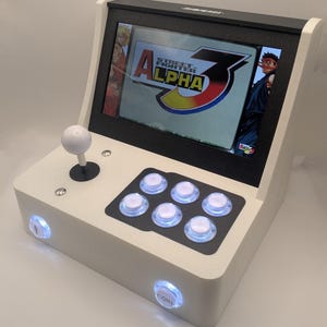 May include: A white and black mini arcade machine with a screen showing "Street Fighter Alpha 3". It has a joystick, six action buttons, and two coin buttons. The cabinet has a retro design.