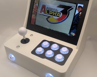 Op de afbeelding: Een wit en zwart mini-arcademachine met een scherm met "Street Fighter Alpha 3". Het heeft een joystick, zes actieknoppen en twee muntknoppen. De kast heeft een retro-ontwerp.