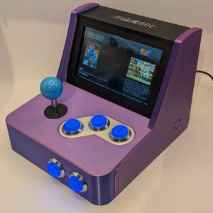 Può includere: Una console arcade da tavolo viola etichettata Minicade con uno schermo che mostra l'interfaccia del gioco Neo-Geo Metal Slug 2. Il dispositivo presenta un joystick blu, cinque pulsanti circolari blu con bordi argentati e un pannello superiore nero.