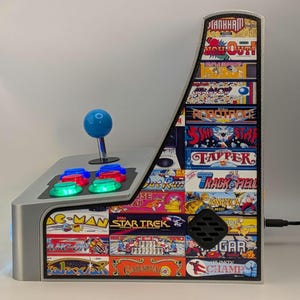May include: A silver tabletop arcade game with a blue joystick and colorful buttons. The cabinet features classic arcade game titles like Pac-Man and Star Trek. The side panel displays various game art and the text "Tankham" and "Out!"