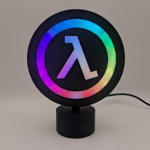以下が含まれることがあります： 中央に白いラムダ記号が付いた黒い円形LEDランプ。シンボルはカラフルな光のリングで囲まれています。ランプは黒い円筒形のベースに取り付けられており、黒い電源コードが付いています。