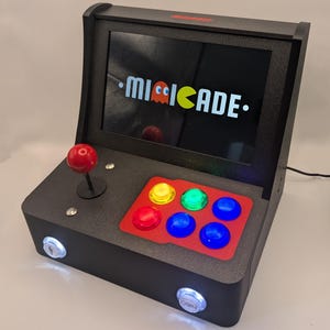 Könnte beinhalten: Schwarze Mi-Cade Arcade-Maschine mit einem Bildschirm, der das Mi-Cade-Logo anzeigt. Sie hat einen roten Joystick, mehrfarbige Knöpfe und zwei Münzschlitze. Die Maschine ist für Spiele konzipiert.