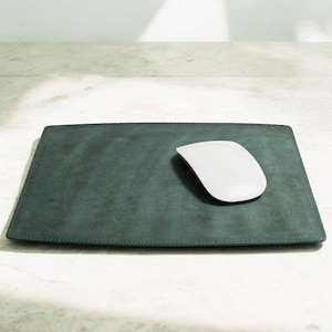 May include: A rectangular, dark teal leather mousepad with a white wireless mouse. The mousepad has stitched edges and sits on a light-colored surface. The mouse is positioned on the right side of the pad.