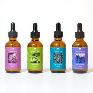 Könnte beinhalten: Vier braune Glasflaschen mit schwarzen Tropfern und farbigen Etiketten. Die Etiketten lauten "Lilac Flower", "Balsam Cedar", "Balsam Fir" und "Lavender Balsam". Jedes Etikett trägt auch die Aufschrift "Car Freshener Refill".