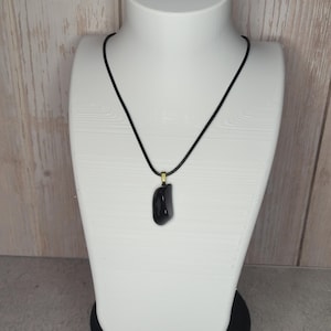 Op de afbeelding: Een zwarte stenen hangerketting, tentoongesteld op een witte buste. De hanger is een gladde, onregelmatig gevormde zwarte steen, hangend aan een zwart koord met een goudkleurige sluiting. De buste staat tegen een lichte houtnerf achtergrond.