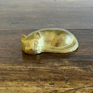 May include: A brass cat ornament in a resting pose. The cat has etched features and a textured surface. The figurine is golden in colour and rests on a wooden surface.