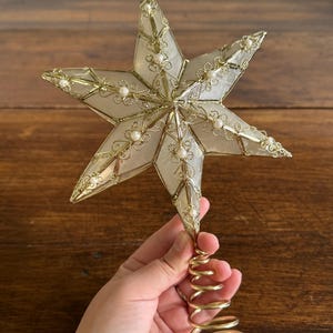 May include: A six-pointed star tree topper featuring translucent white panels framed in gold-toned wire filigree. The star is accented with small white beads along the edges and includes a gold-toned spiral coil base for attachment to a tree.