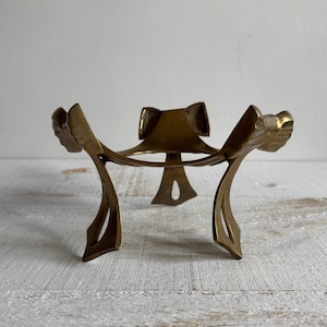 Könnte beinhalten: Ein dekorativer Vintage-Ständer aus Messing mit einem einzigartigen, durchbrochenen Design. Der Ständer hat ein zentrales bogenförmiges Element und gebogene Stützen. Das Messing hat eine warme, gealterte Patina, was darauf hindeutet, dass es sich um ein antikes oder Vintage-Stück handelt. Der Ständer ist für ein kleines Objekt konzipiert.