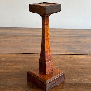 May include: A tall, dark brown wooden pedestal stand featuring a square base, a slender tapered central column, and a flat square top platform. The wood grain is visible throughout the structure, showcasing a polished, stained finish.