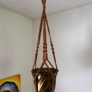 May include: A brown macrame plant hanger featuring knotted cord construction with a top loop for suspension, holding a metallic bronze-coloured planter with a spiraled, fluted design and a smooth, reflective surface finish.