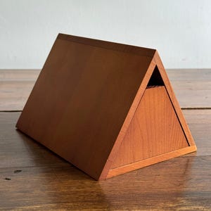 May include: A triangular wooden box with a smooth, medium-brown finish. The object features a hinged lid that forms one of the slanted sides of the prism shape, with a visible seam along the top edge and a triangular side panel.