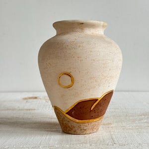 May include: A small, beige ceramic vase with a textured surface. The vase features a brown and gold geometric design near the base and a gold circle near the top. The vase has a rounded body and a narrow neck.
