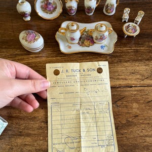 May include: A collection of miniature porcelain tea set pieces featuring gold trim and floral scenes, including a teapot, sugar bowl, tray, pitchers, and chairs, alongside a vintage J. R. Tuck & Son receipt from Port Colborne, Ontario, dated July 7, 1972.