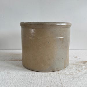 May include: A cylindrical stoneware crock featuring a smooth, light beige finish with subtle variations in tone. The vessel has a thick, rounded rim at the top and a straight-sided body with a slightly textured surface, showing a simple, functional design.