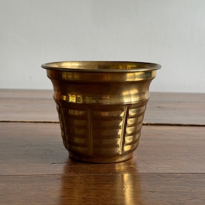 May include: A small, polished brass planter with a flared rim and a cylindrical body featuring vertical panels and horizontal ribbed detailing. The metallic surface reflects light, highlighting the warm golden tones and the textured design.