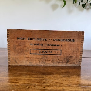 May include: A distressed wooden box, marked with the text "HIGH EXPLOSIVE -- DANGEROUS" and "CLASS III -- DIVISION I" in black. The box also features "C.R.C. 14". The box is brown and rests on a wooden surface.