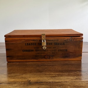 May include: A rectangular brown wooden ammunition crate with a hinged lid and a brass latch secured by a small padlock. The front features black printed text reading LOADED SHOT GUN SHELLS, MADE IN CANADA BY CANADIAN INDUSTRIES LIMITED, MONTREAL, CANADA.