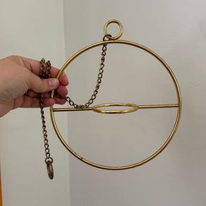 Peut inclure: Un support de plante suspendu circulaire en métal doré, composé d'un grand anneau extérieur, d'un anneau central plus petit pour un pot et d'une barre de support horizontale. La structure est suspendue par une chaîne en métal doré assortie.