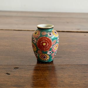 Puede incluir: Un jarrón de cerámica en miniatura con un fondo verde azulado que presenta un patrón denso de flores multicolores en tonos rojos, amarillos, rosas y blancos. El pequeño recipiente tiene un cuerpo redondeado, cuello estrecho y acabado brillante.