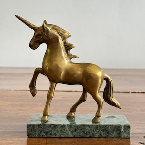 May include: A brass unicorn figurine standing in a prancing pose with one front leg raised. The mythical creature features a pointed horn, a flowing mane, and a tail. It is mounted on a rectangular, polished green marble base with visible veining.