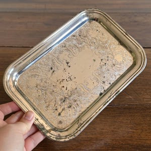 May include: A rectangular silver-toned metal tray featuring an ornate embossed scrollwork pattern along the border. The surface displays visible signs of tarnish and dark oxidation spots throughout the centre and edges of the metallic finish.